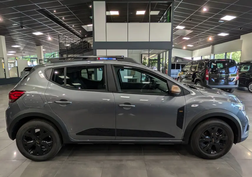 Vue de profil droit d'une Dacia Sandero Stepway TCe 110 gris schiste avec jantes noires en intérieur de showroom.