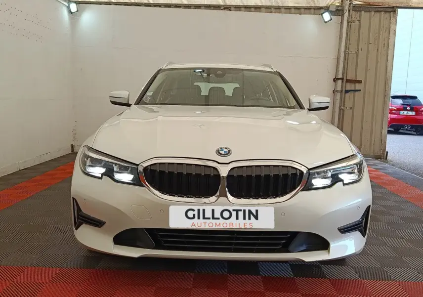 Vue avant d'une BMW Série 3 320e blanche avec calandre noire et phares LED allumés dans un garage.