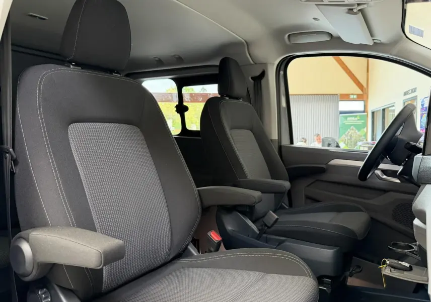 Vue intérieure côté gauche du Volkswagen Transporter T7 ProCab 2026, sièges tissu gris à rayures avec accoudoirs.