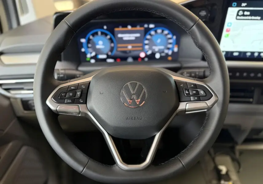 Volant multifonction noir du Volkswagen Transporter T7 avec tableau de bord numérique et écran tactile visible en arrière-plan.