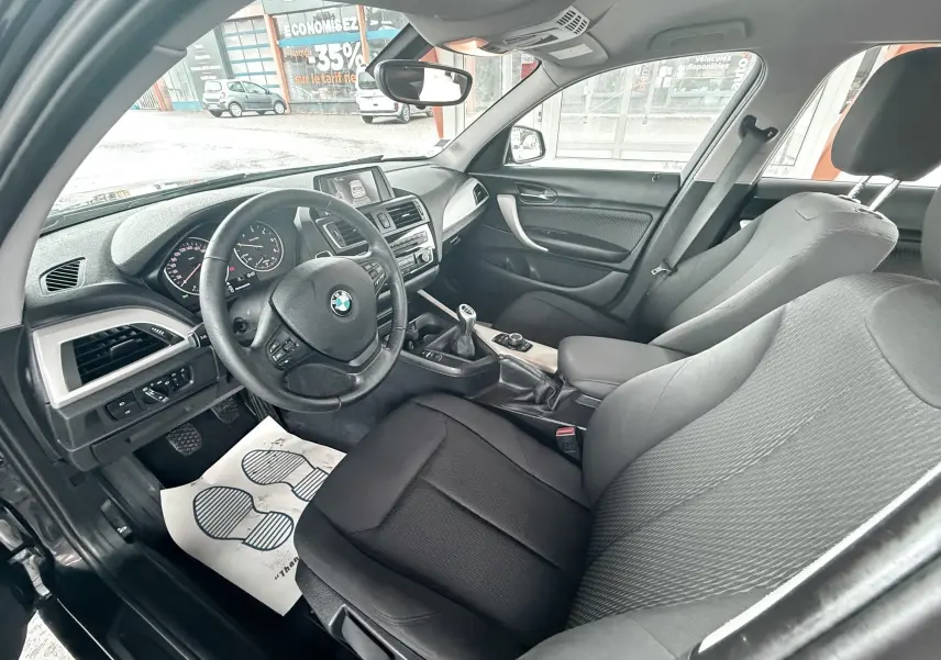Vue intérieure côté conducteur de la BMW Série 1 noire, montrant le volant, la boîte manuelle et les sièges en tissu noir.