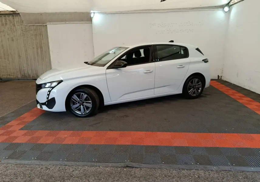 Peugeot 308 blanche vue de profil côté gauche, dans un espace fermé avec sol gris et bandes rouges.