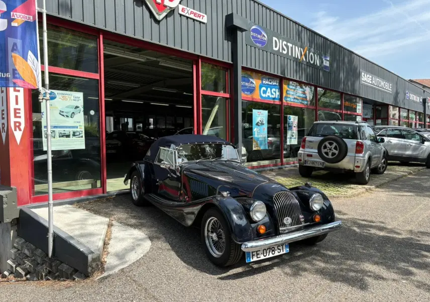 Morgan Plus 4 bleu foncé vue 3/4 avant droit, capote fermée, garée devant un garage avec façade vitrée rouge.