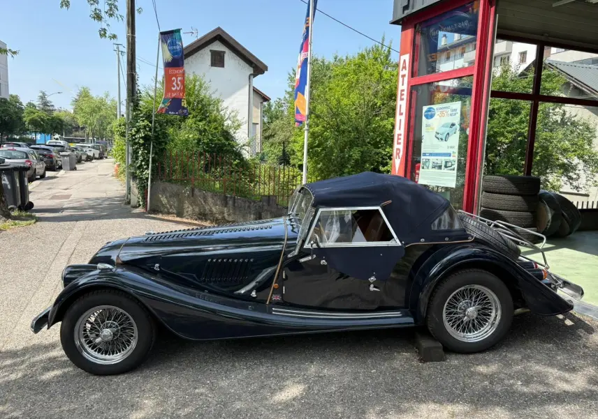 Morgan Plus 4 bleu foncé vue de profil côté gauche, capote fermée et jantes à rayons chromées visibles.
