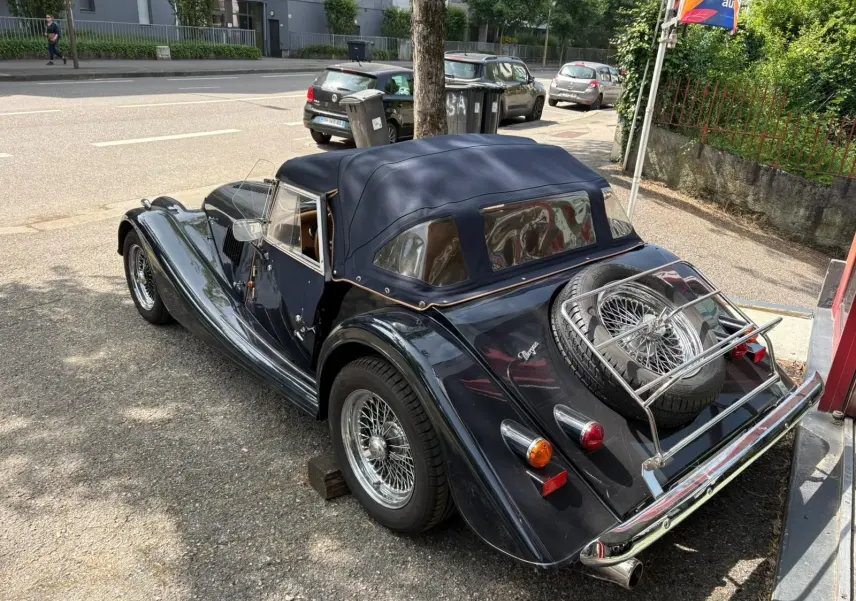 Morgan Plus 4 bleu foncé vue 3/4 arrière droit, capote fermée et roue de secours sur coffre chromé.