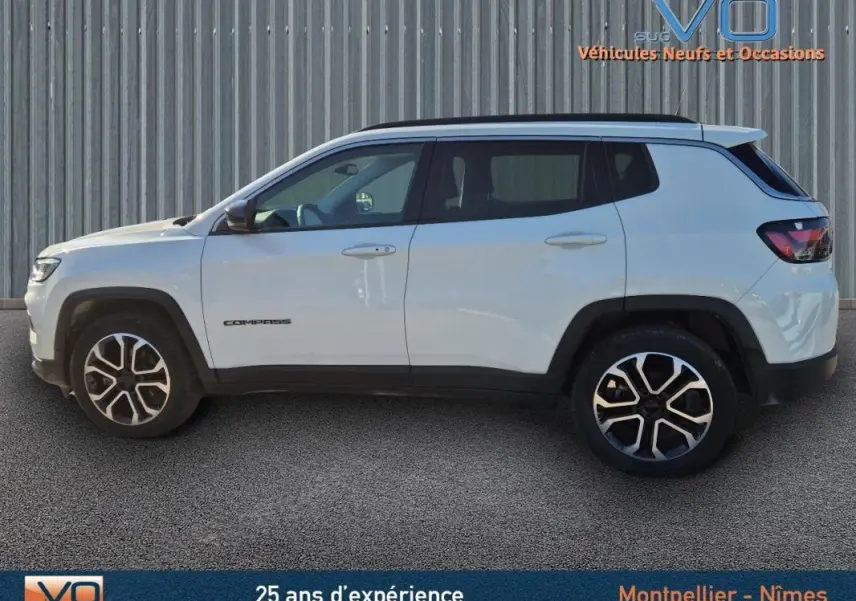 Profil latéral droit d'un Jeep Compass blanc 2023 avec jantes 18 pouces et barres de toit visibles sur fond industriel.