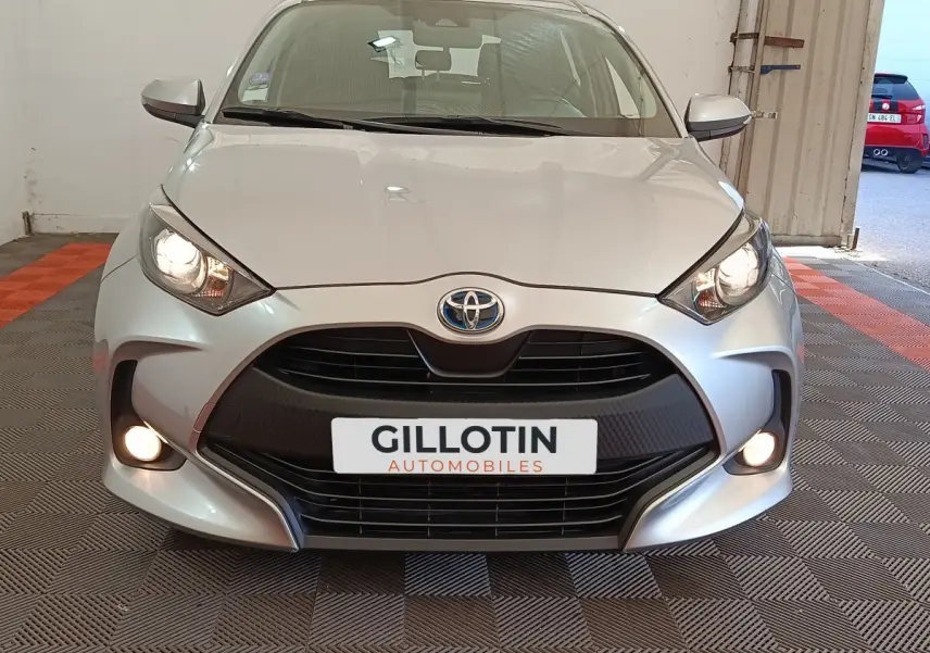 Vue frontale d'une Toyota Yaris hybride gris clair avec phares et antibrouillards allumés en intérieur.