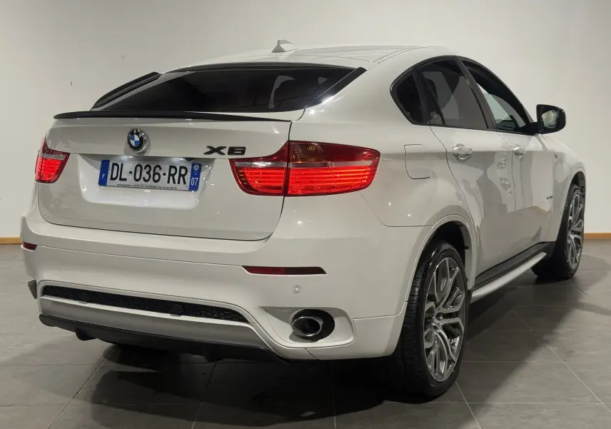 Vue 3/4 arrière droite d’un BMW X6 blanc 2026 avec feux allumés et jantes M Sport distinctives.