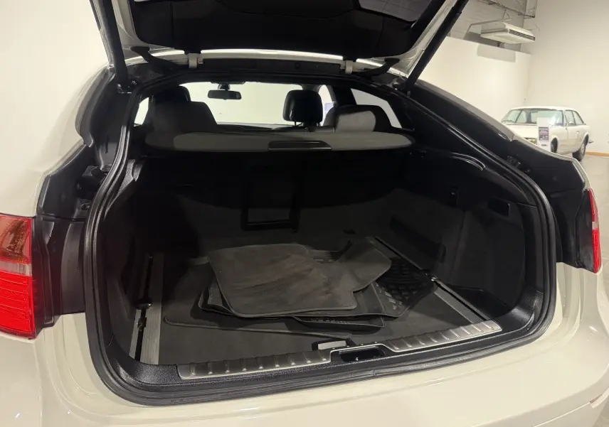 Coffre ouvert vu de l'arrière d'un BMW X6 blanc 2026 avec tapis de sol noirs à l'intérieur.