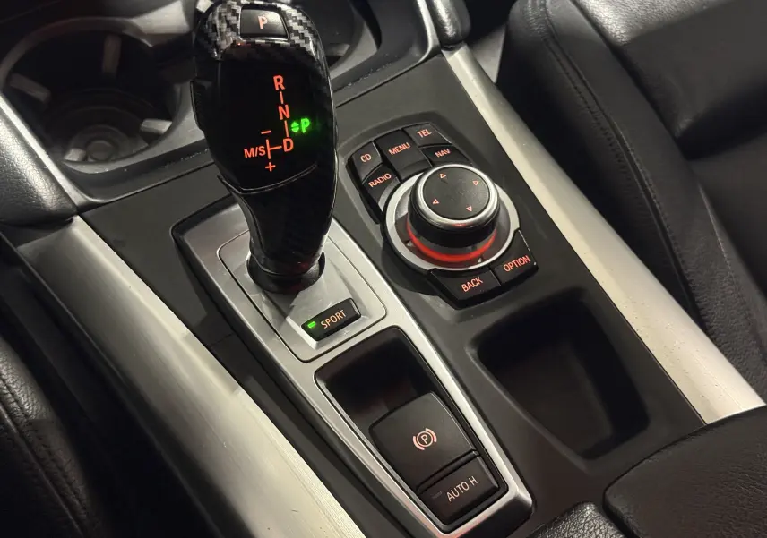 Gros plan sur la console centrale du BMW X6 blanc 2026, avec levier de vitesses carbone et commandes iDrive éclairées.