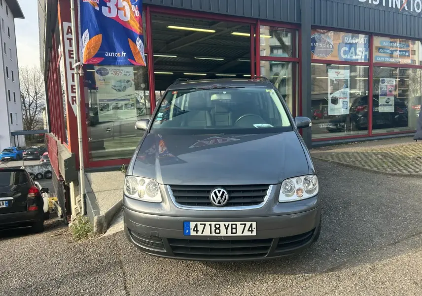 Volkswagen Touran gris 2004 vu de face, stationné devant un atelier avec une bannière publicitaire visible.