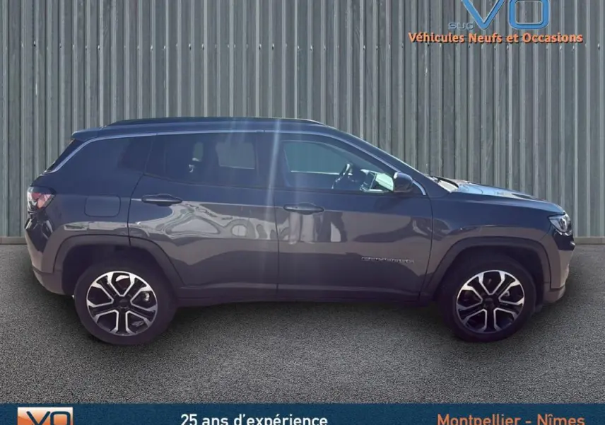 Vue latérale droite d'un Jeep Compass gris 2023 avec jantes 18 pouces et barres de toit visibles sur fond industriel.