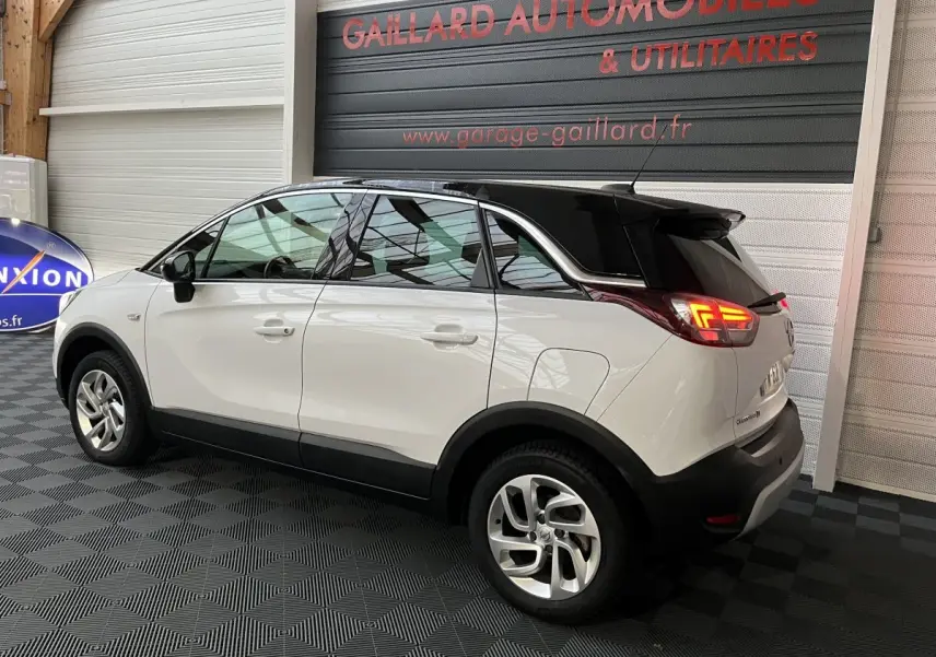 Vue 3/4 arrière droite d'un Opel Crossland X blanc avec toit noir, feux arrière allumés, stationné en intérieur.