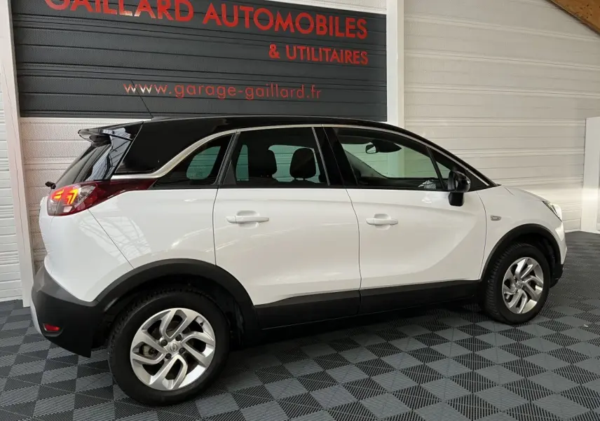 Opel Crossland X blanc vue de profil côté gauche, toit noir contrastant et feux arrière allumés en intérieur showroom.