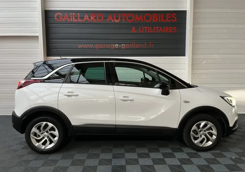 Vue de profil côté gauche d'un Opel Crossland X blanc avec toit noir, roues alliage et protections noires basses.