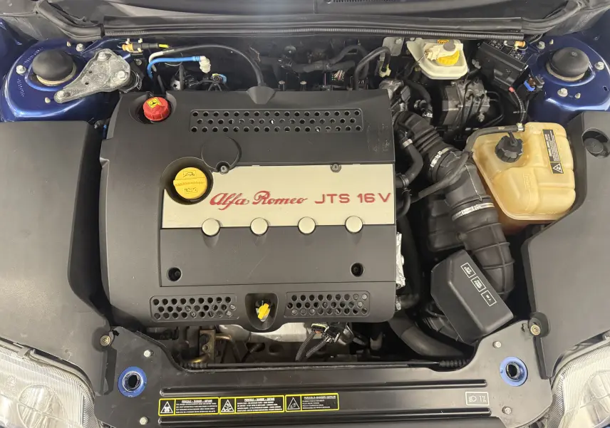 Vue plongeante sur le moteur noir et gris marqué Alfa Romeo JTS 16V d’une Alfa Romeo Spider bleu 2006.