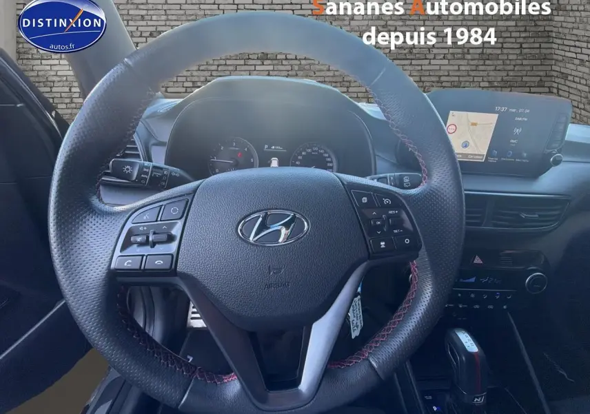 Vue intérieure centrée sur le volant cuir N Line avec surpiqûres rouges et écran tactile du Hyundai Tucson gris foncé.