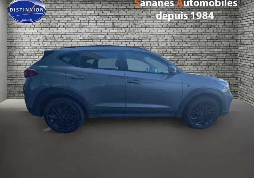 Vue latérale droite du Hyundai Tucson 1.6 Hybrid 136 N Line gris foncé avec jantes alliage noires, sur fond de mur en briques.
