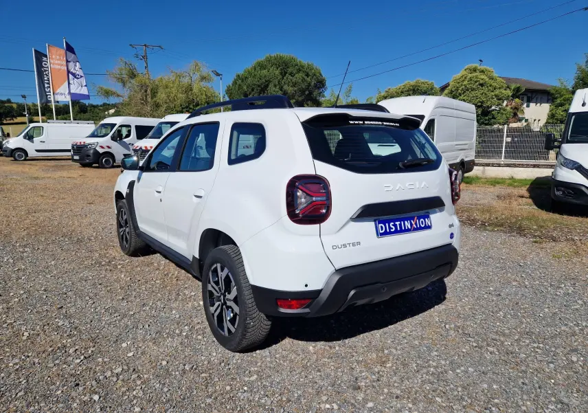 Vue 3/4 arrière droite d'un Dacia Duster blanc 2024 avec jantes alu 17 pouces, sur un parking extérieur ensoleillé.