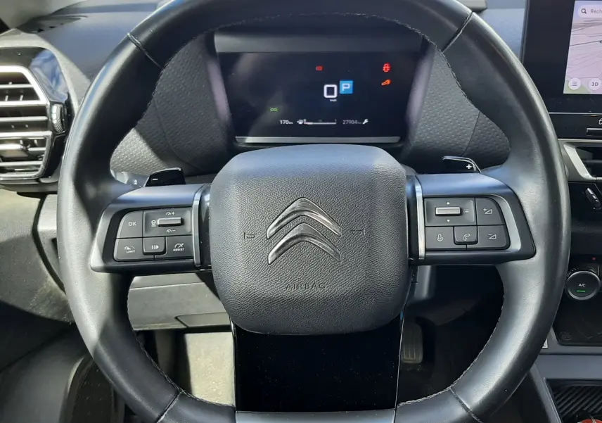 Vue rapprochée du volant noir de la Citroën C4 2024 avec commandes intégrées et tableau de bord numérique en arrière-plan.