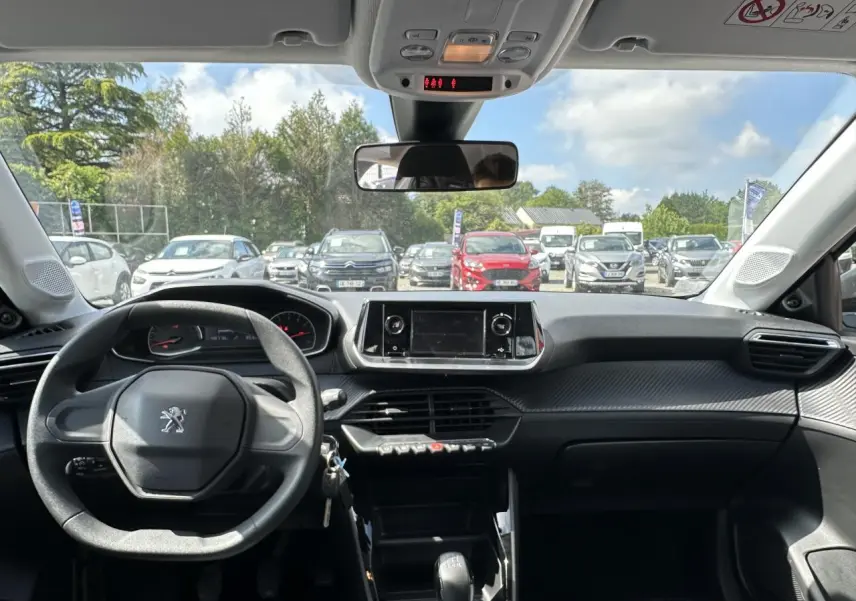 Vue intérieure avant du tableau de bord noir et volant Peugeot 208 blanc, avec écran tactile et ciel bleu visible à l'extérieur.