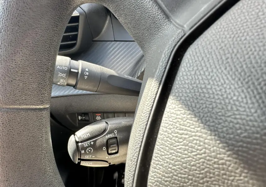 Gros plan sur la commande de régulateur de vitesse à gauche du volant d’une Peugeot 208 blanche, intérieur noir.