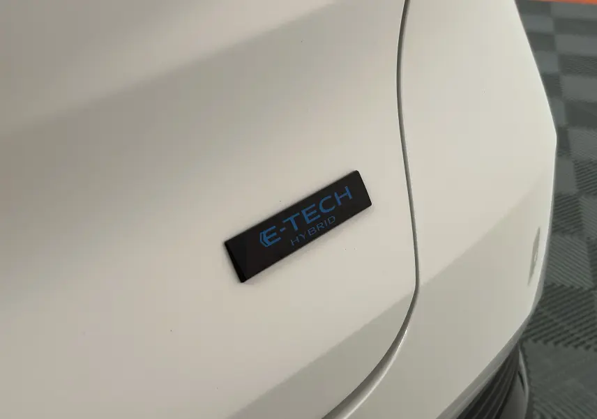 Gros plan sur l'emblème E-TECH Hybrid bleu sur la carrosserie blanc nacré d'un Renault Captur II Ph.2.