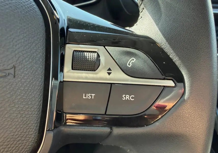 Gros plan sur les commandes noires et argentées du volant côté gauche d’une Peugeot 208 gris Artense 2025.