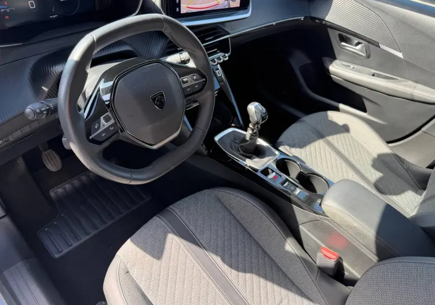 Vue intérieure côté conducteur de la Peugeot 208 1.2 100ch S&S Allure avec volant compact, sièges tissu gris et levier de vitesse manuel.