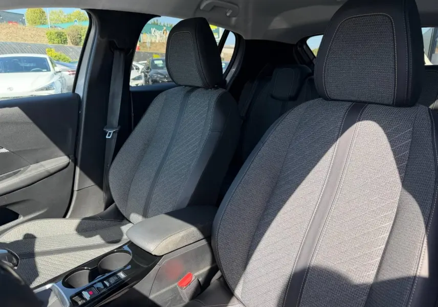 Intérieur de la Peugeot 208 2025, vue côté avant droit sur sièges tissu gris avec accoudoir central noir.