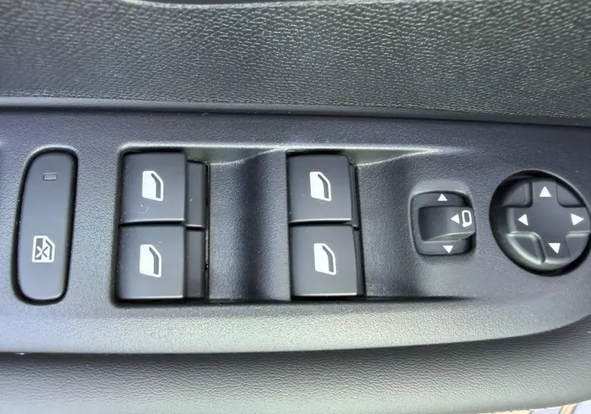 Gros plan sur les commandes électriques des vitres et réglages des rétroviseurs côté conducteur d'une Peugeot 208 gris Artense.