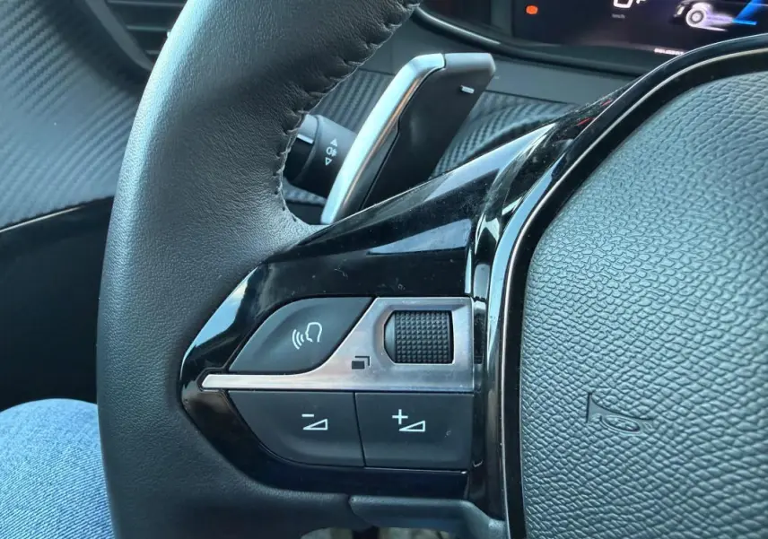 Gros plan sur le côté gauche du volant noir de la Peugeot 208 Hybrid 110 e-DCS6 Allure avec commandes audio et palette de vitesse.