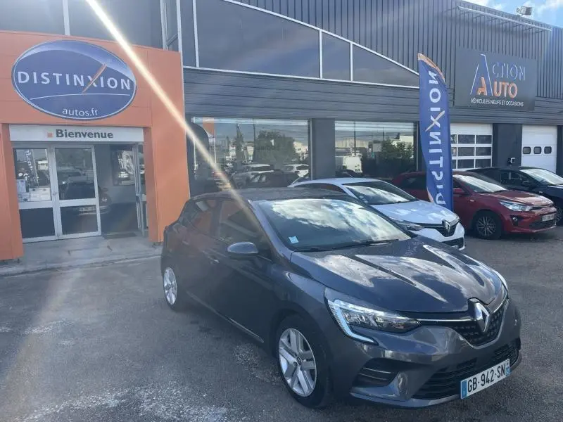 Renault Clio gris titanium vue 3/4 avant droit devant un concessionnaire avec drapeau Distinxion visible