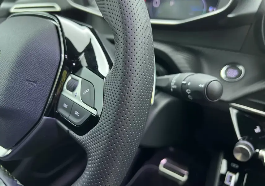 Gros plan sur le volant perforé et les commandes intégrées du tableau de bord noir de la Peugeot 208 1.2 Hybrid 136 GT.