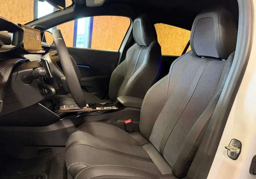 Vue intérieure côté conducteur de la Peugeot 208 blanche 2025, mettant en valeur les sièges en tissu noir et le tableau de bord moderne.