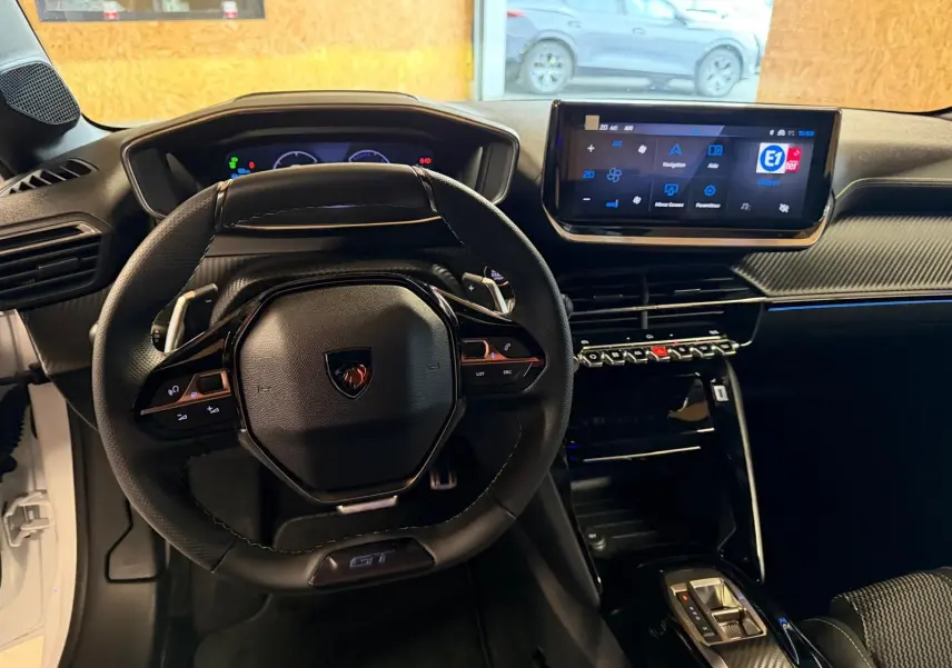 Vue intérieure du poste de conduite de la Peugeot 208 1.2 HYBRID 136 GT blanche, avec volant compact et écran tactile central.
