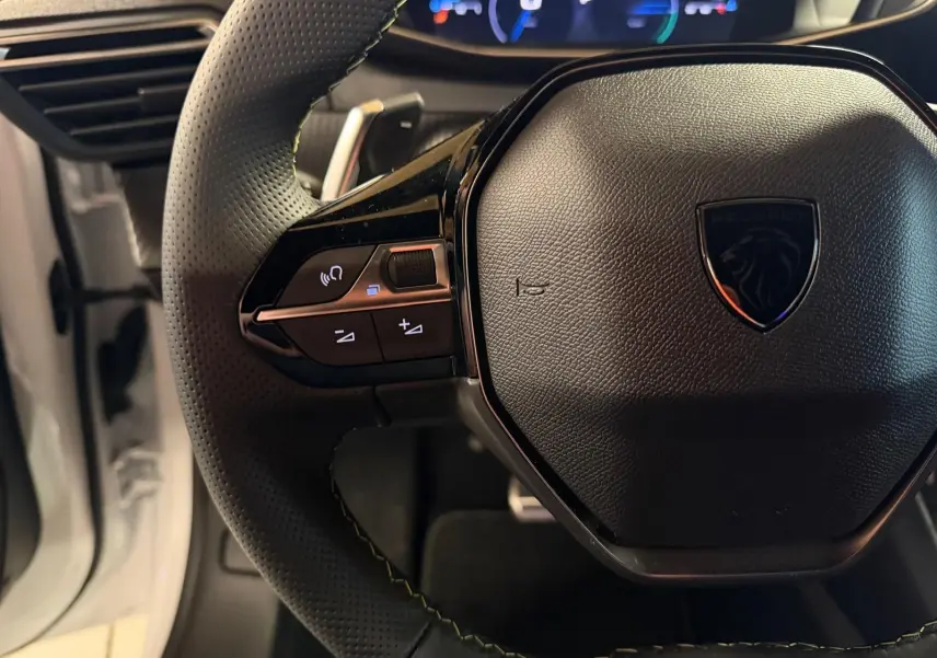 Gros plan sur le volant noir perforé du Peugeot 208 1.2 HYBRID 136 GT blanc avec commandes intégrées et tableau de bord digital.