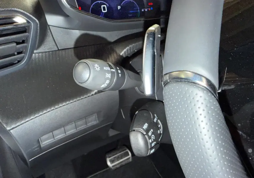 Gros plan sur la partie gauche du volant perforé et les commandes de feux et régulateur d’une Peugeot 208 blanche hybride.