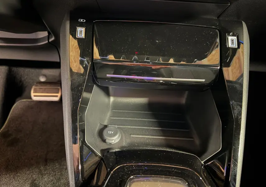Vue rapprochée de la console centrale noire brillante du Peugeot 208 1.2 Hybrid 136 GT avec prise 12V visible.