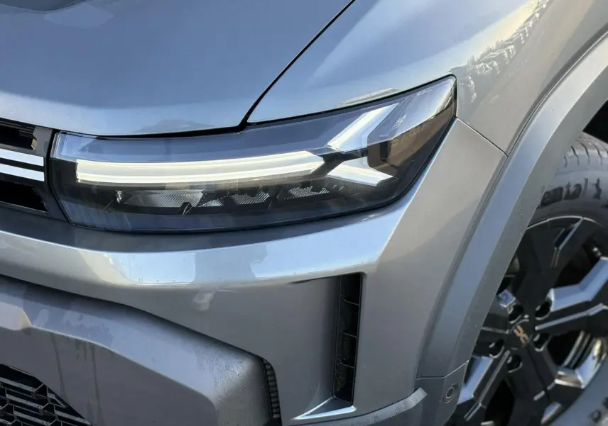 Gros plan sur le phare avant droit et la calandre du Dacia Duster gris schiste avec jante alliage noire visible.