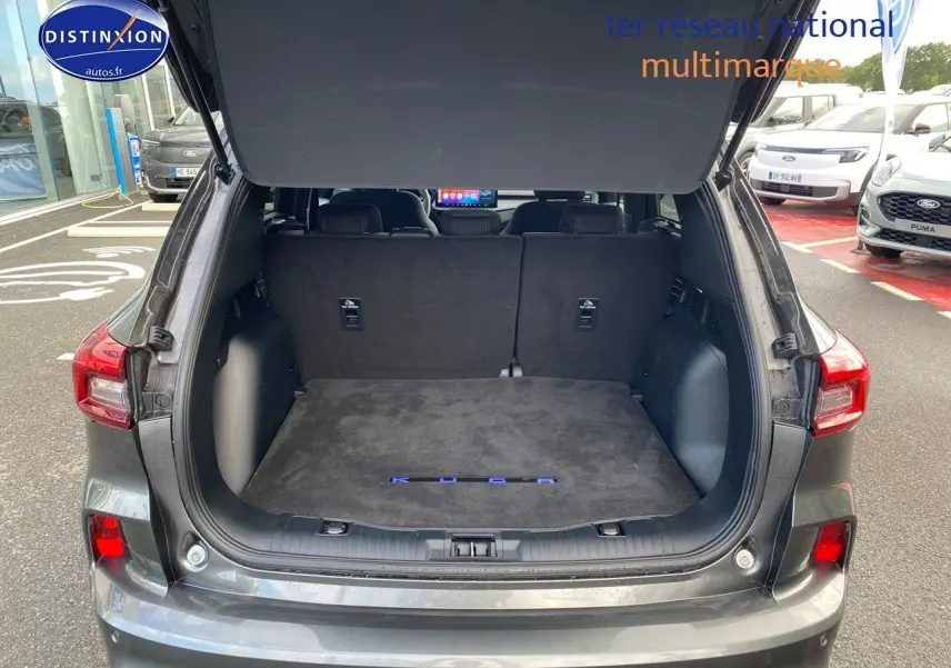 Coffre ouvert en vue arrière d’un Ford Kuga gris Magnetic Fashion Métallisée avec tapis de coffre noir marqué Kuga.