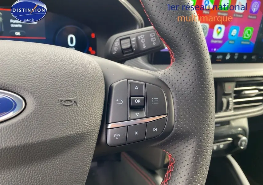 Gros plan sur le volant perforé avec surpiqûres rouges du Ford Kuga 2025, affichage numérique et écran tactile en arrière-plan.