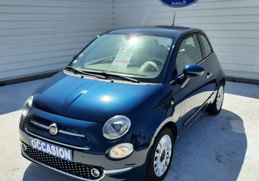 Vue 3/4 avant d'une FIAT 500 bleu foncé avec phares ronds et jantes blanches, stationnée devant un mur blanc.