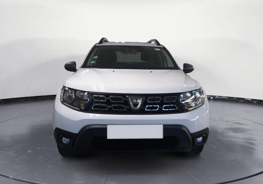 Dacia Duster blanc en 3/4 avant droit, avec calandre noire et jantes alliage 16 pouces, stationné sur un sol gravillonné.