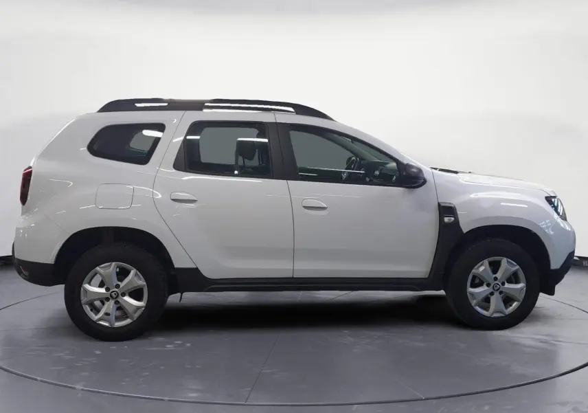 Vue 3/4 arrière droite d'un Dacia Duster blanc 2021 avec jantes alliage et barres de toit sous un abri.