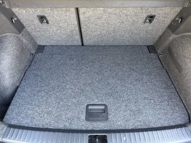 Coffre arrière vu de face, revêtement gris clair, avec poignée d'accès au rangement sous plancher.