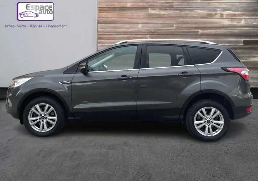 Profil côté gauche d'un Ford Kuga gris 2017 avec jantes alliage et barres de toit, garé devant un mur en bois.