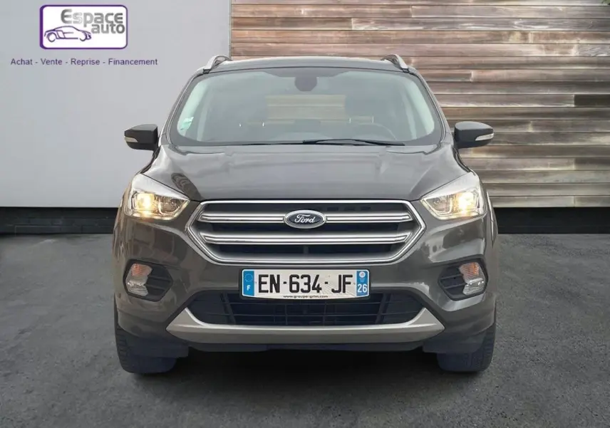 Vue frontale d'un Ford Kuga gris 2017 avec phares allumés et calandre chromée, stationné devant un mur en bois.