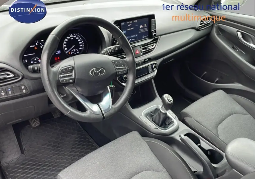 Vue intérieure de face du poste de conduite de la Hyundai i30 SW blanche, volant multifonction et boîte manuelle visibles.