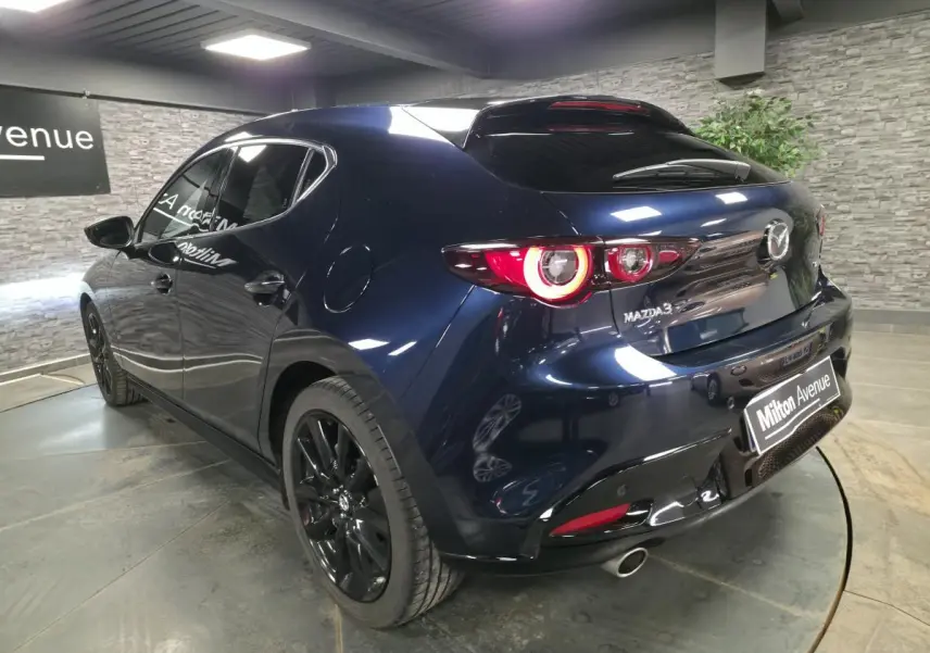 Vue 3/4 arrière droite d'une Mazda3 2024 bleu métallisé avec jantes noires et feux arrière LED circulaires allumés.